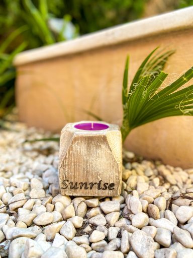 Portavelas de madera con la palabra "Sunrise" y fondo de piedras y plantas verdes.