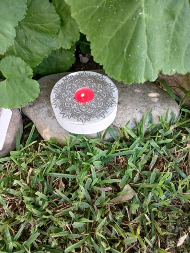 Una vela blanca con un toque rojo sobre una piedra, rodeada de hierba y hojas verdes.
