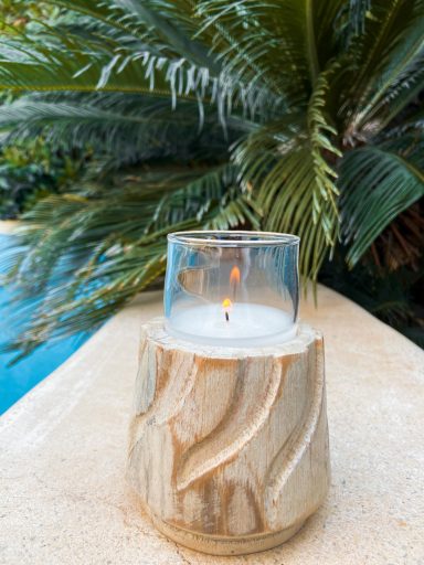 Vela encendida sobre un soporte de madera, junto a una piscina y plantas tropicales.