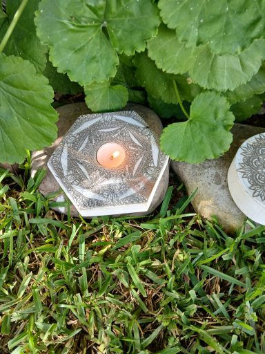 Una vela encendida sobre una piedra hexagonal, rodeada de hojas verdes y césped.