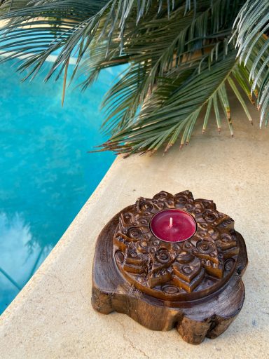 Portavelas decorativo de madera con vela roja, junto a una piscina y palmeras.